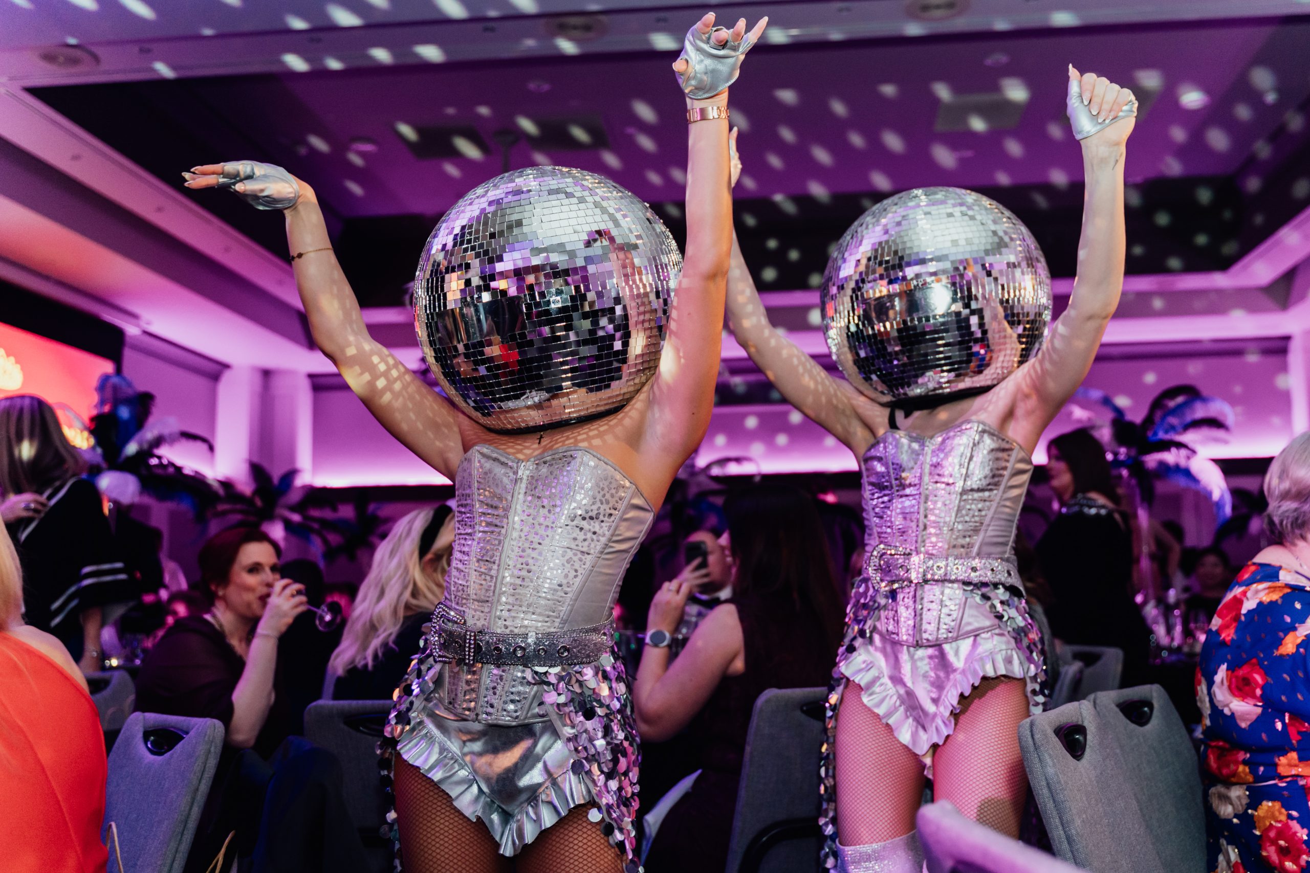 Purple lit ball room of awards attendees with two dancers wearing disco ball heads move through the crowd